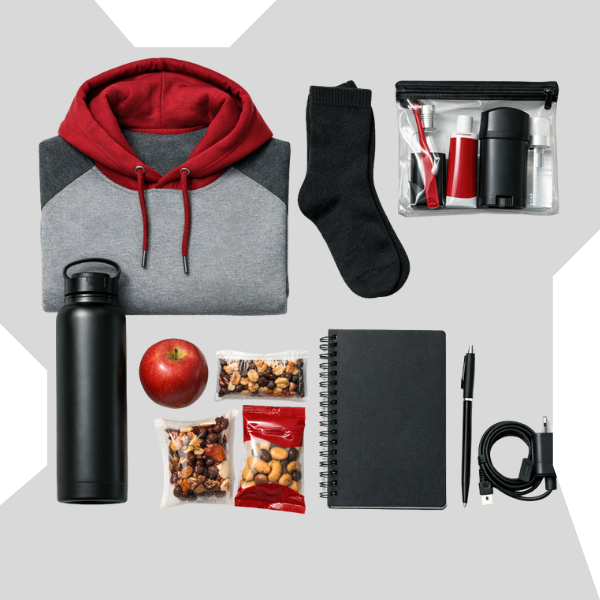 Flat-lay of basic needs care package items for teens including a hoodie, socks, hygiene kit, water bottle, snacks, notebook, pen, and phone charger, arranged on a neutral background.
