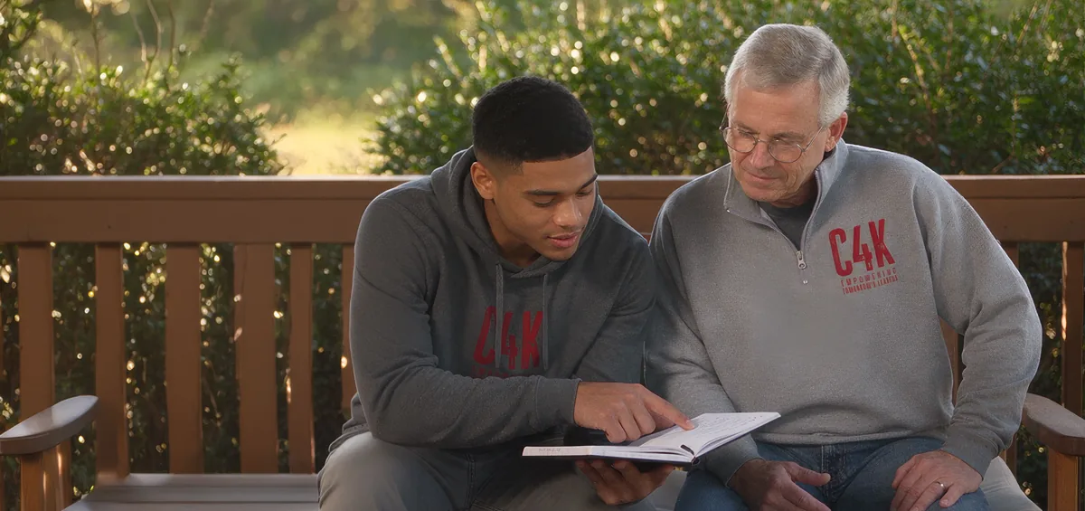 A college man and an alumnus in C4K apparel reviewing notes on an outdoor bench